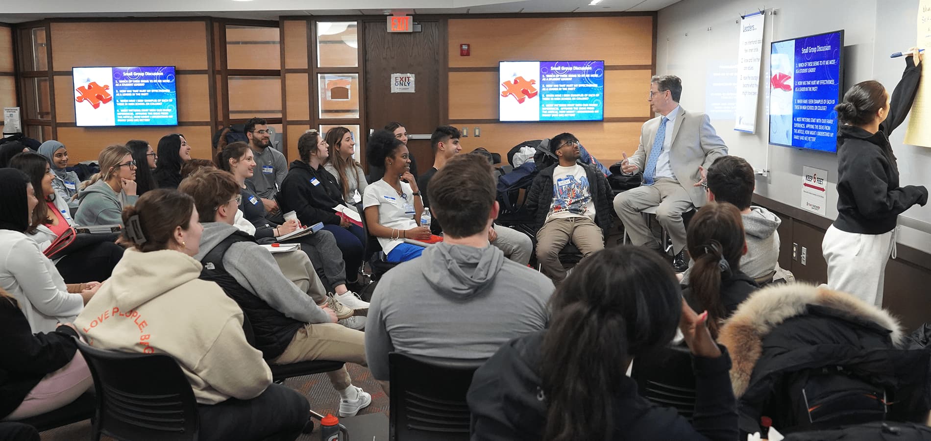 Students sit inside of a classroom and listen to a teacher who sits on a higher chair, with three television screens around them.