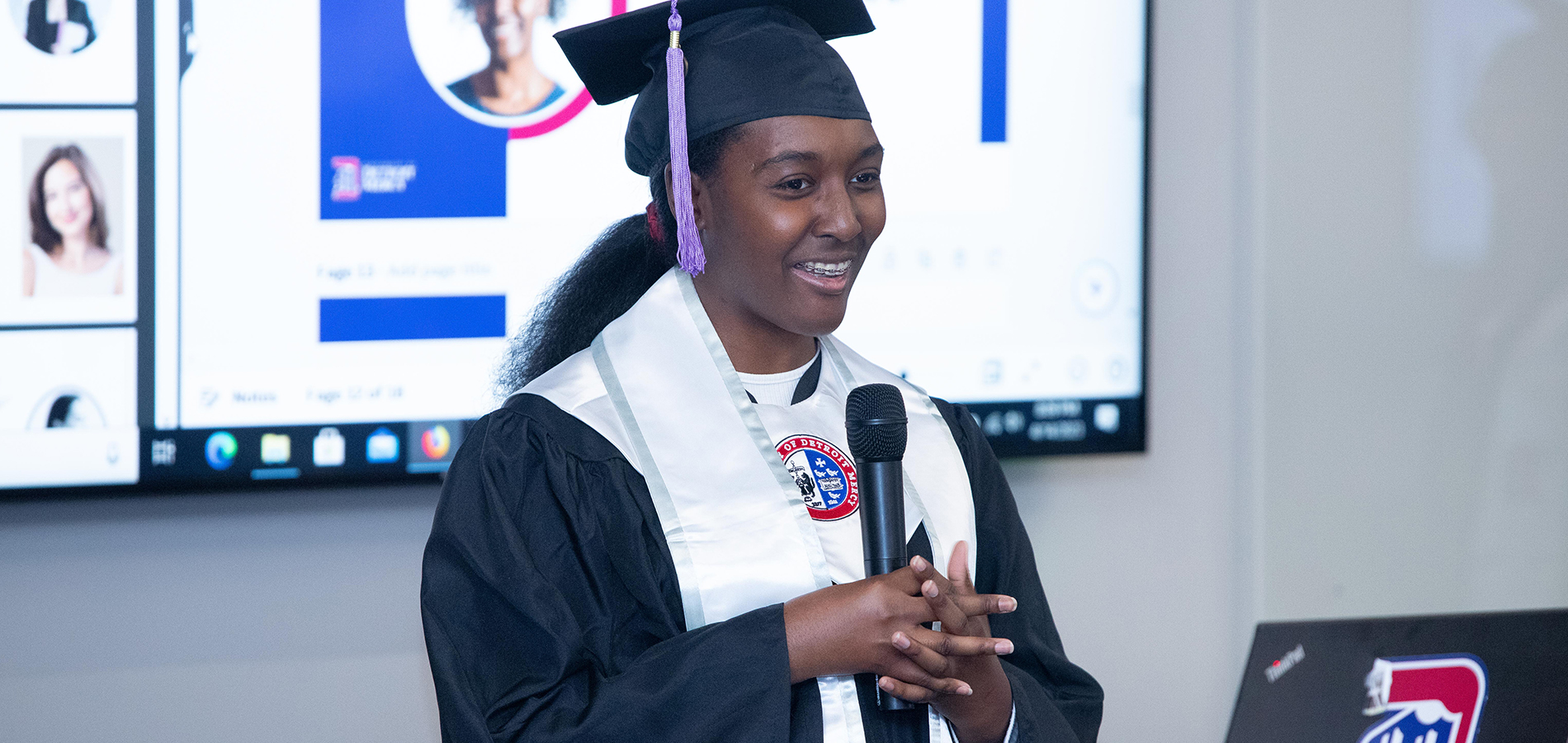 Angel Mangham wears a graduation cap and gown and speaks from a podium inside of a classroom.