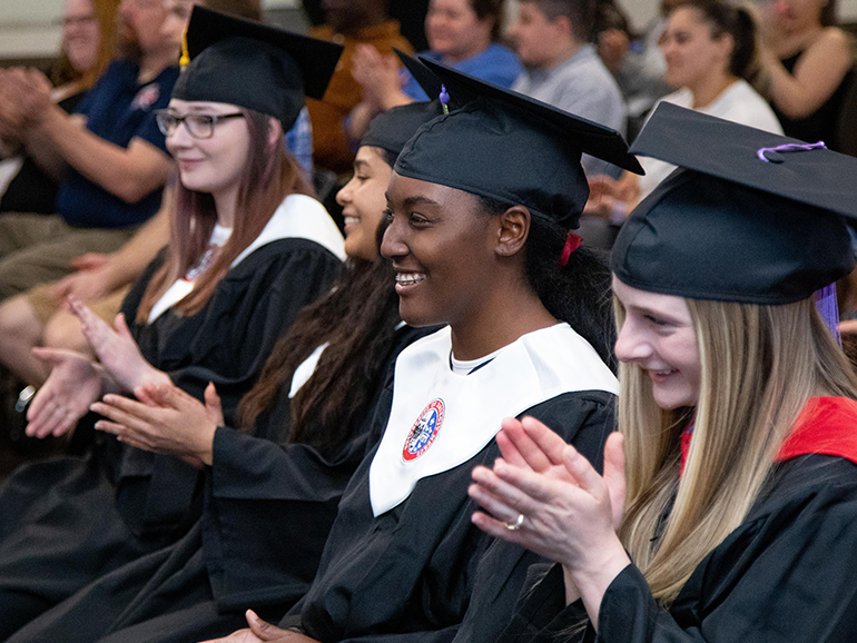 Angel Mangham sits and smiles with other graduates and people sitting inside of a classroom.