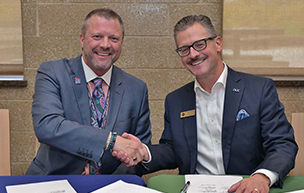Two people sit at a table and shake hands, while smiling for the camera inside of a building.