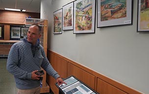 Brian Owen points to the display for an exhibit of paintings owned by his family. The paintings, hanging on the wall and behind showcase glass, are part of the McNichols Campus Library's latest exhibit.