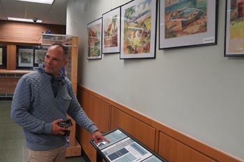 Brian Owen points to the display for an exhibit of paintings owned by his family. The paintings, hanging on the wall and behind showcase glass, are part of the McNichols Campus Library's latest exhibit.