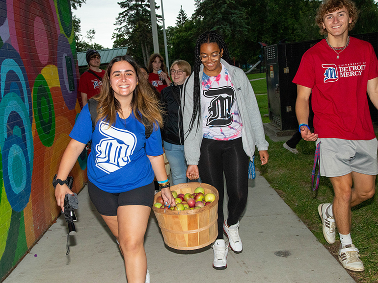 Three students smile wearing University of Detroit Mercy t-shirts, with two of them carrying a bucket of apples outdoors. Other students are pictured behind them and a colorful brick wall is next to them.
