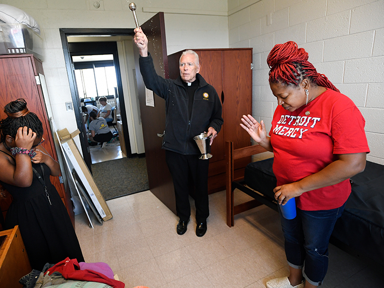 A father blesses a room with two people standing inside of it. One wears a red Detroit Mercy t-shirt. Across the hallway others can be seen in a dorm room of Shiple Hall.