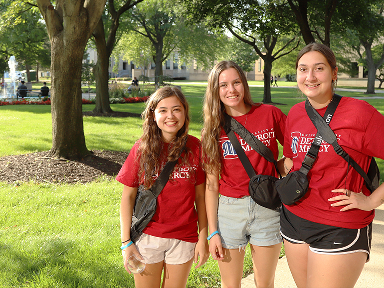 Three students wearing red University of Detroit Mercy t-shirts smile for a photo during a sunny day outdoors on the McNichols Campus.