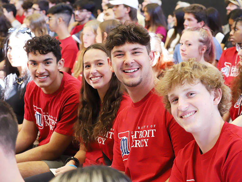 Four students wearing red University of Detroit Mercy t-shirts smile for a photo while sitting indoors. Other students are pictured behind them.