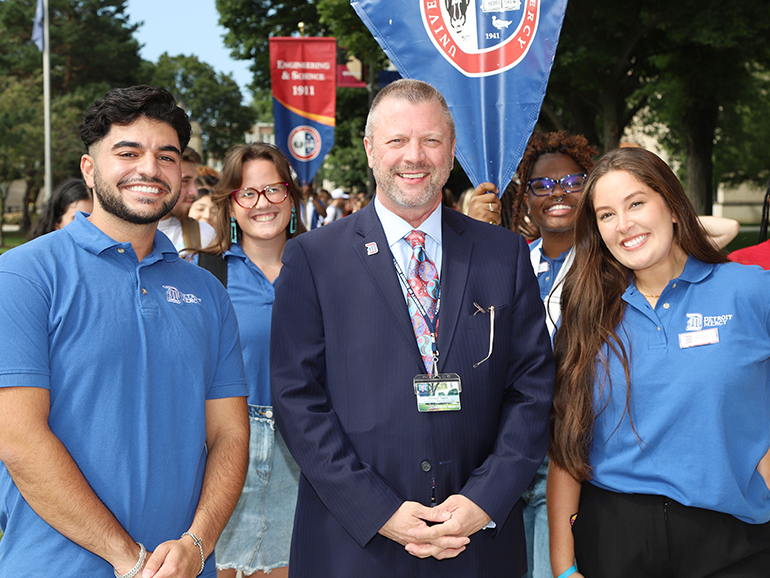Four student leaders pose for a photo with President Taylor, with others behind outdoors on the McNichols Campus.