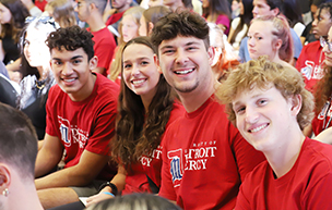 Four students wearing red University of Detroit Mercy t-shirts sit and smile for a photo indoors, with other students in the background.