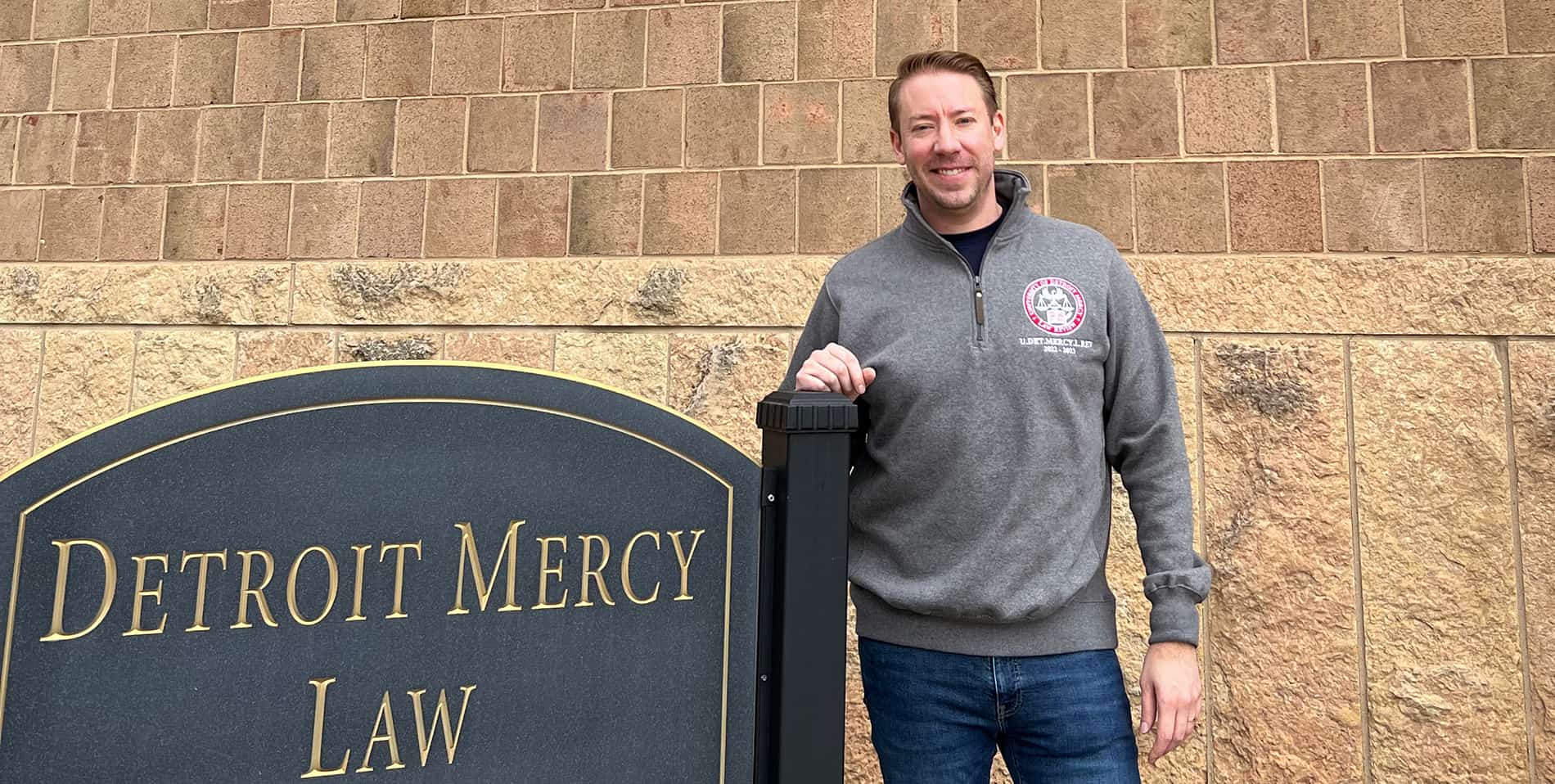 Matthew Snyder stands in front of the School of Law, next to a sign that reads Detroit Mercy Law.