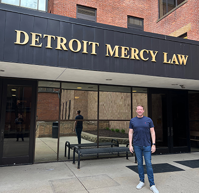 Matthew Snyder stands next to the entrance of the School of Law underneath a sign that reads Detroit Mercy Law.