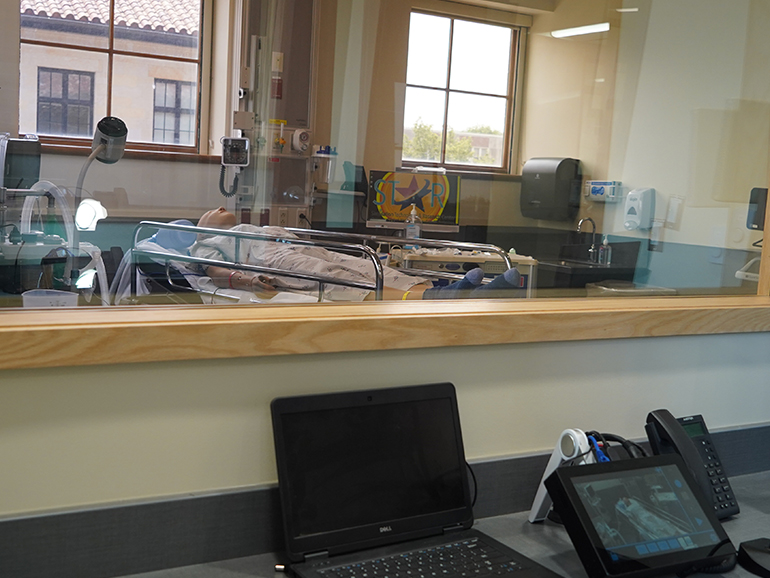 A photo from inside a control room in the STAR Center, looking into a simulation lab classroom with medical equipment.