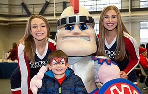 Three people smile for a photo, one a child with his face painted like Spiderman. Tommy Titan is also featured in the center of the photo indoors.