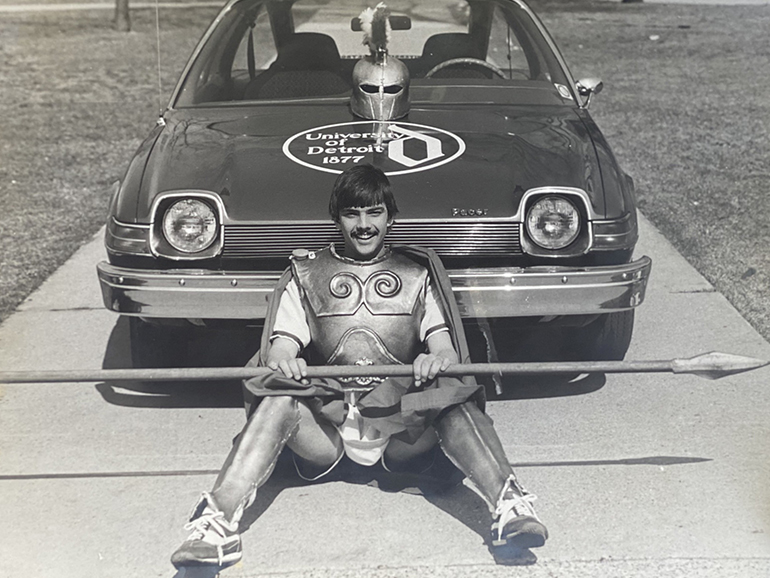 Wearing his mascot outfit, Tom Cieszkowski sits in front of his car with a University of Detroit logo painted on the hood.