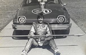 Wearing his mascot outfit, Tom Cieszkowski sits in front of his car with a University of Detroit logo painted on the hood.