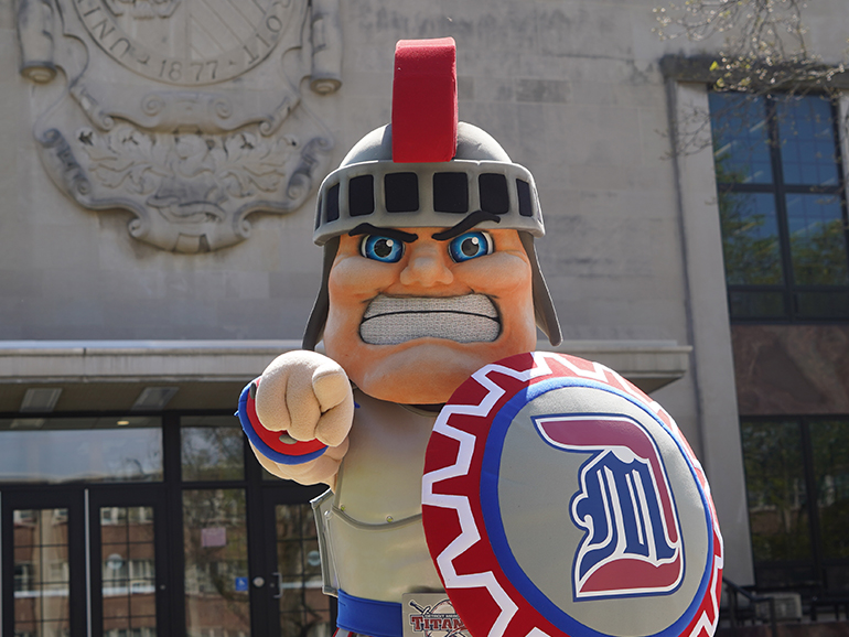 The newest version of Tommy Titan stands in front of the McNichols Campus Library pointing straight ahead and holding his shield.