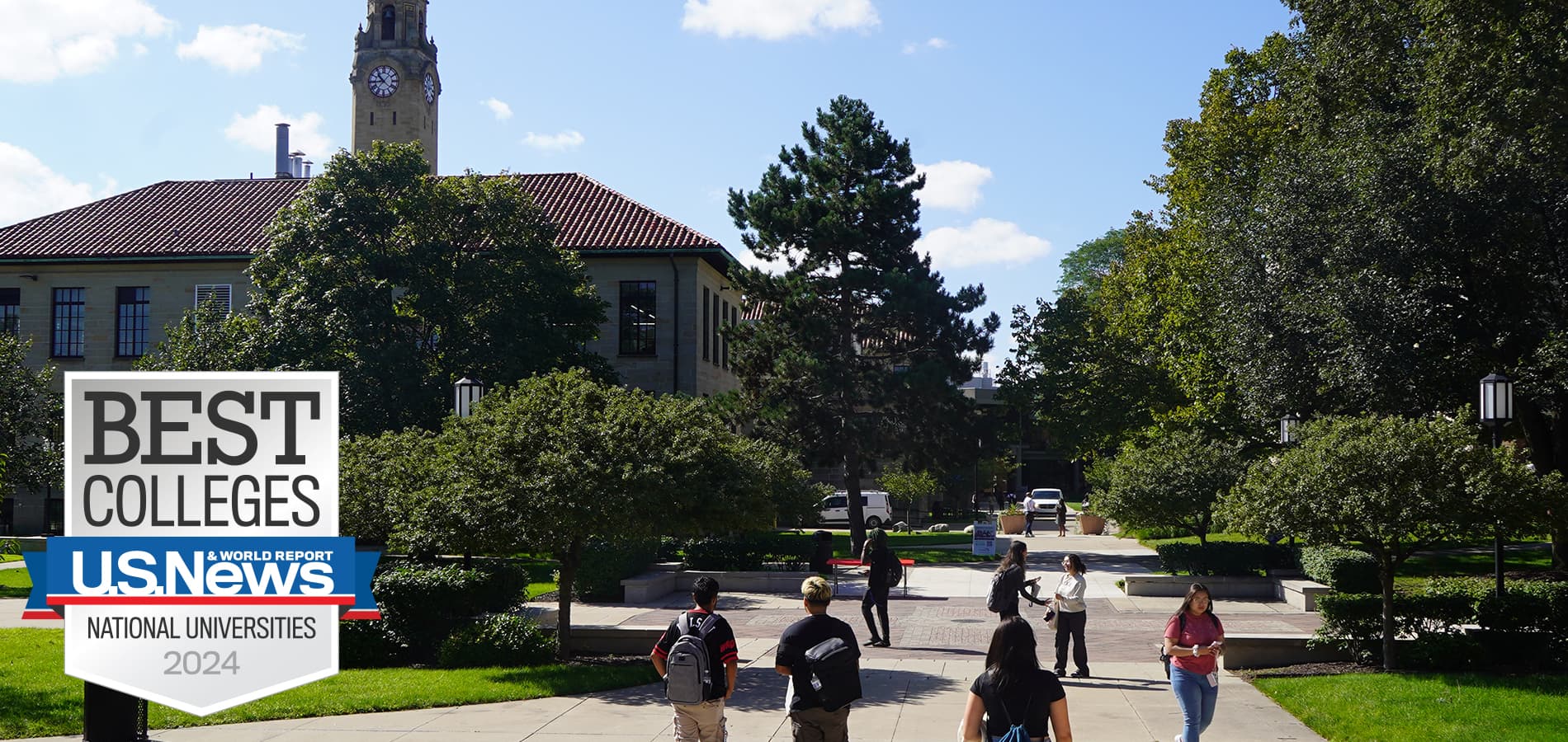 An outdoor photo of the McNichols Campus on a sunny day with students walking on campus amongst trees, buildings and a clock tower.