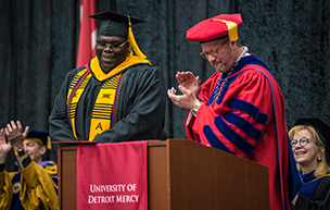 Four people wearing graduation regalia are pictured inside of Calihan Hall during a ceremony. One person claps and another stands at attention next to him. A University of Detroit Mercy red banner is draped over a podium.