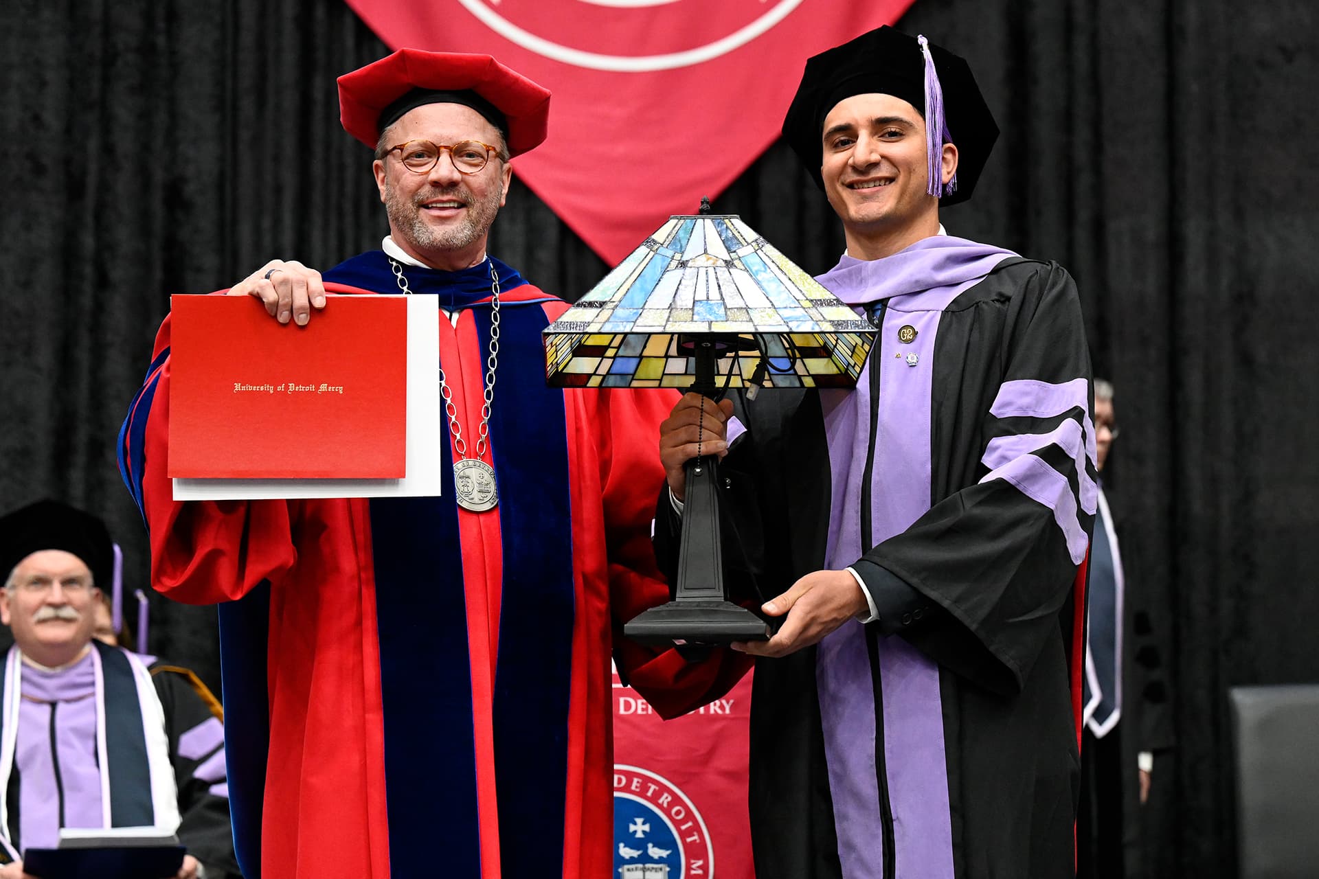 Two people in caps and gowns pose for a photo with a lamp that was presented as an award inside of Calihan Hall.