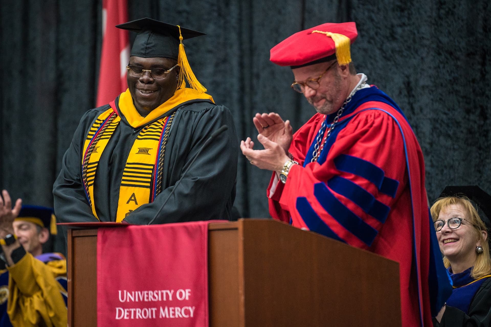 Two people stand on a Calihan Hall stage during a ceremony, one clapping and one smiling. A red University of Detroit Mercy banner hangs from the podium.