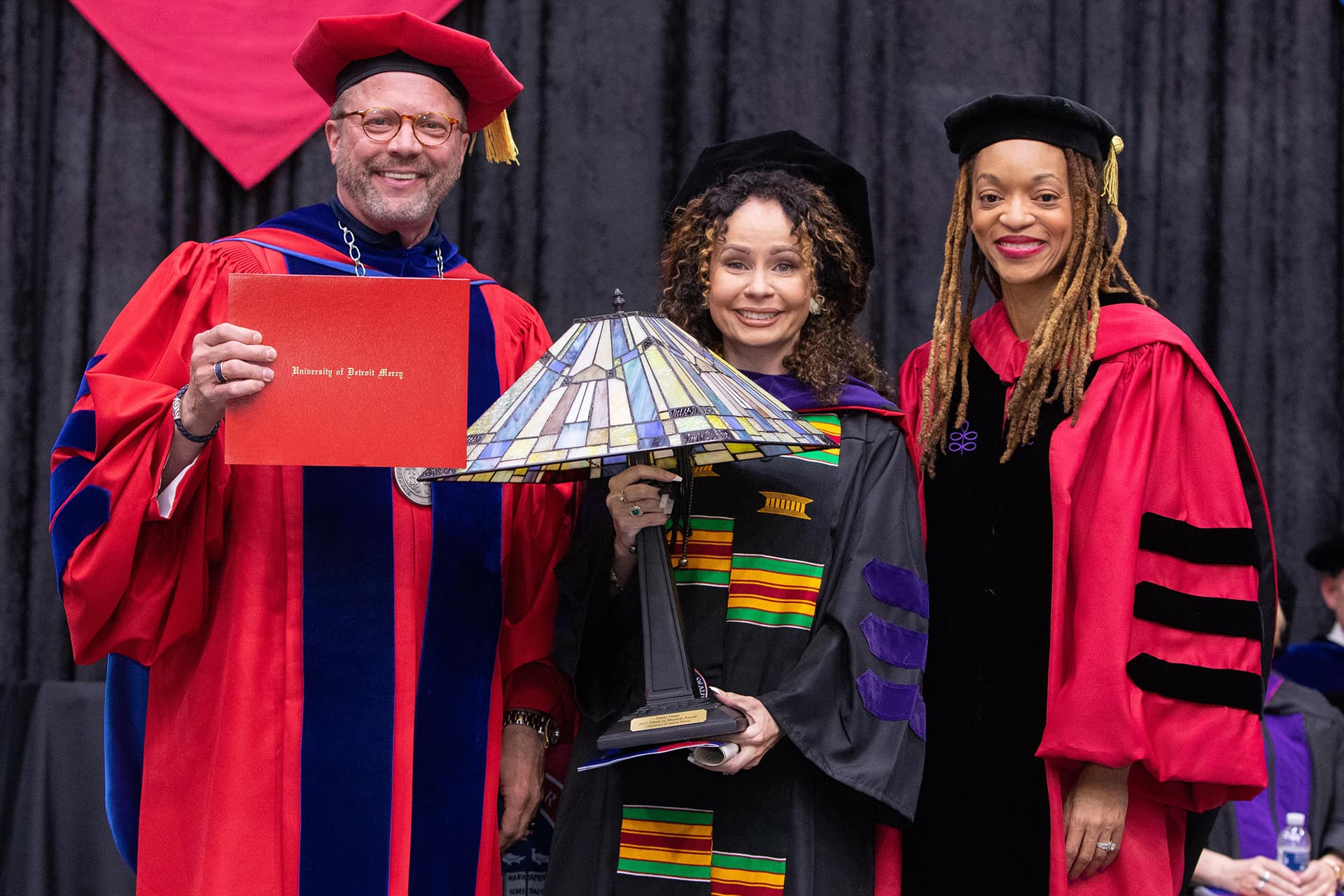 Three people wearing caps and gowns and standing inside of Calihan Hall pose for a photo with a lamp that was presented as an award.