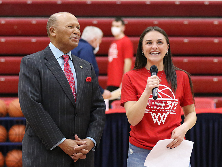 Kara Wolfbauer wearing a Section 313 smiles holding a microphone inside of Calihan Hall, standing next to Dr. Antoine Garibaldi.