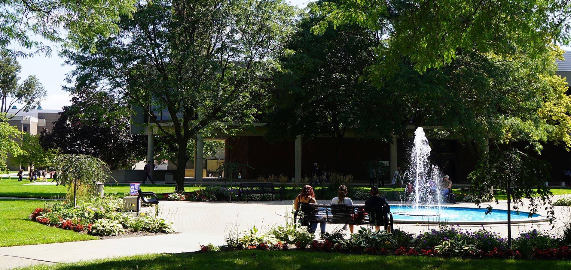 An outdoor photo on the McNichols Campus, featuring students sitting on benches, the Memorial clock tower, water fountain, trees, flowers and buildings during a sunny summer day.