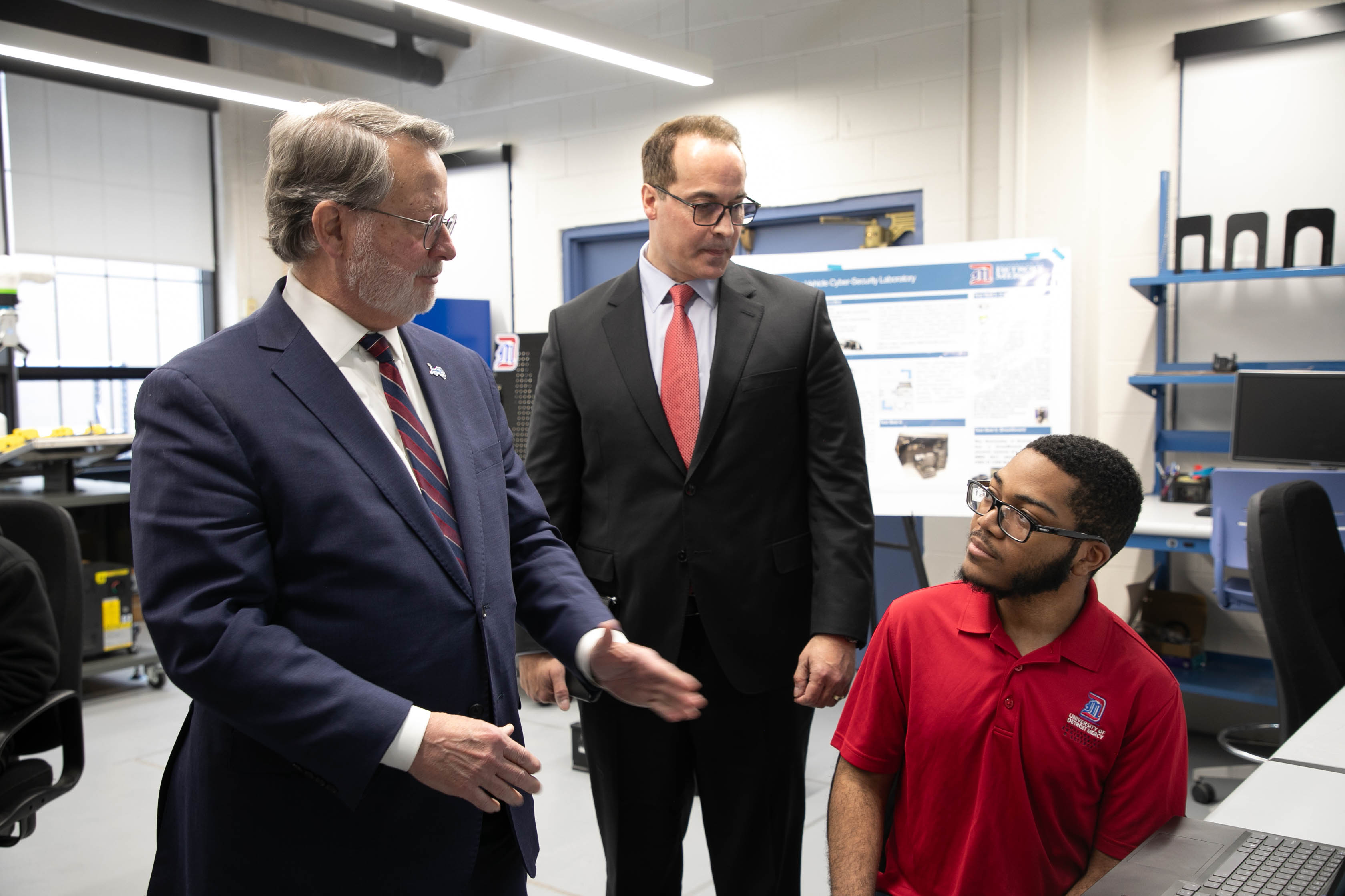 Three people, two standing and one sitting, talk inside of the Engineering Building of Detroit Mercy, with equipment in the background.