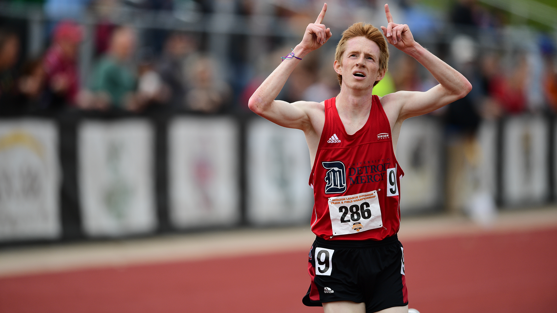 Ben Kendell points to the sky after winning a race outdoors, wearing a red University of Detroit Mercy racing big with numbers on his jersey reading 9 and 286. Logos on his jersey read University of Detroit Mercy and Adidas.