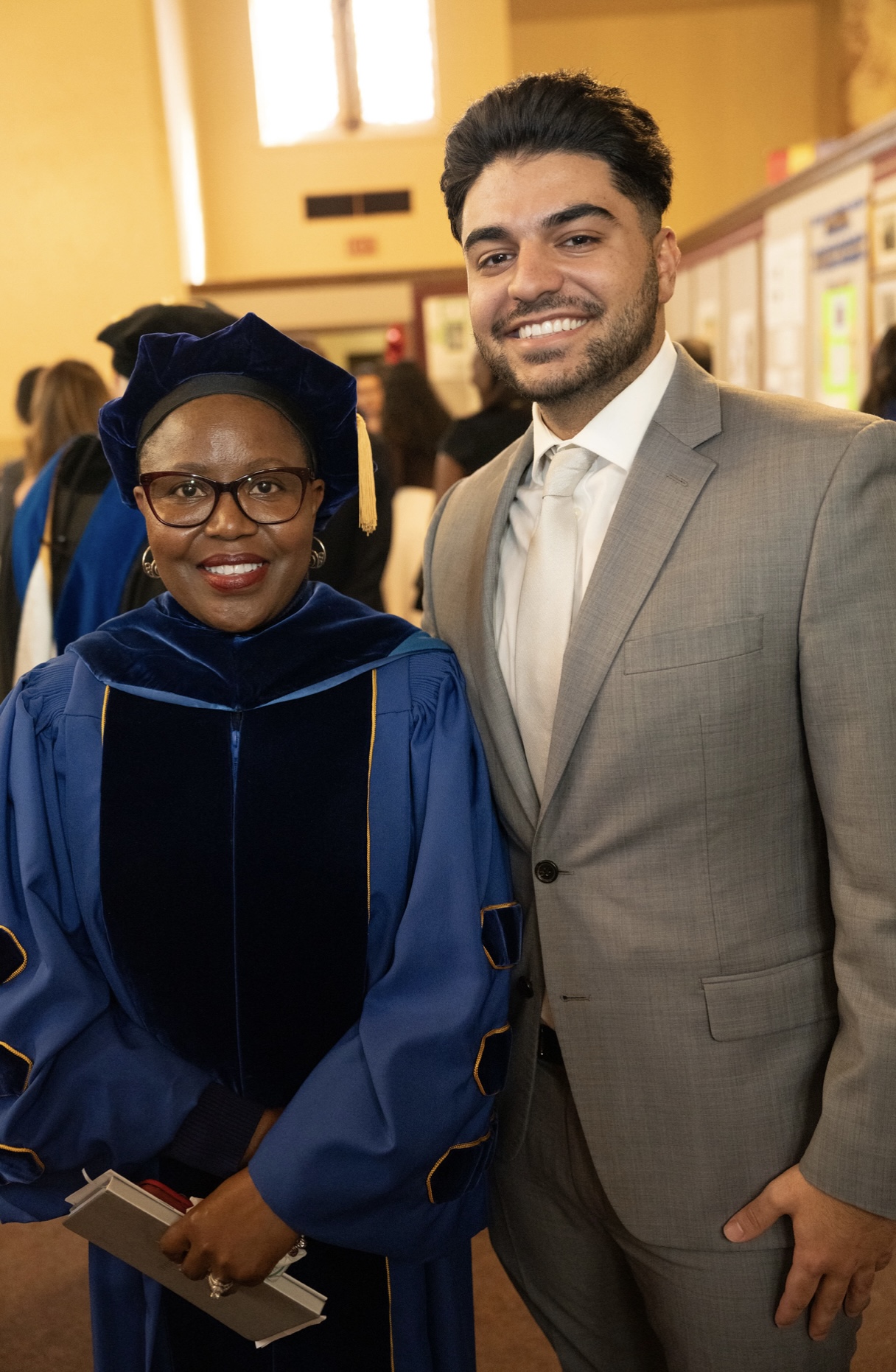 Two people stand indoors posing for a photo, one wearing graduation robes and cap and the other dressed in a suit.