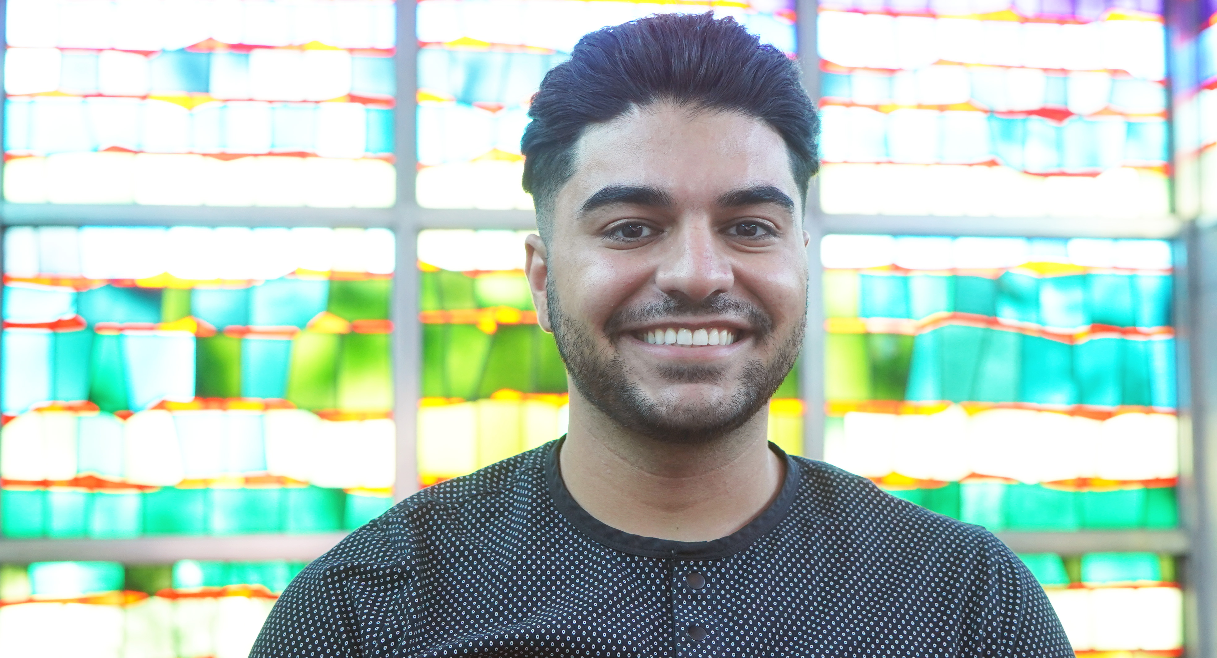 Jacob Yasso stands smiling inside of the Gardella Honors House with stained glass windows behind him.