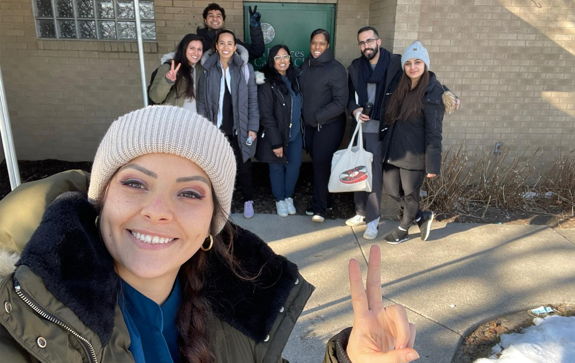 Eight people stand outside of a building, some holding up a peace sign and all smiling.