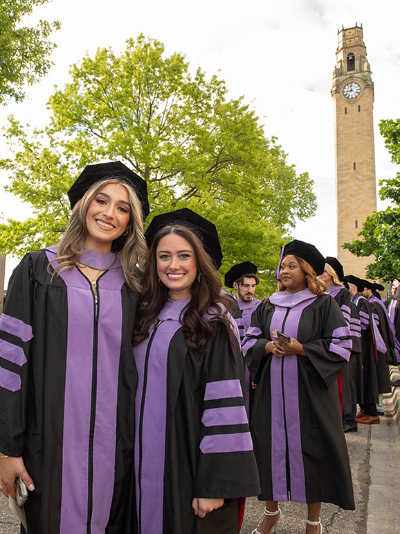 Two graduates take a photo in front of the clocktower as graduates line up for commencement processional.
