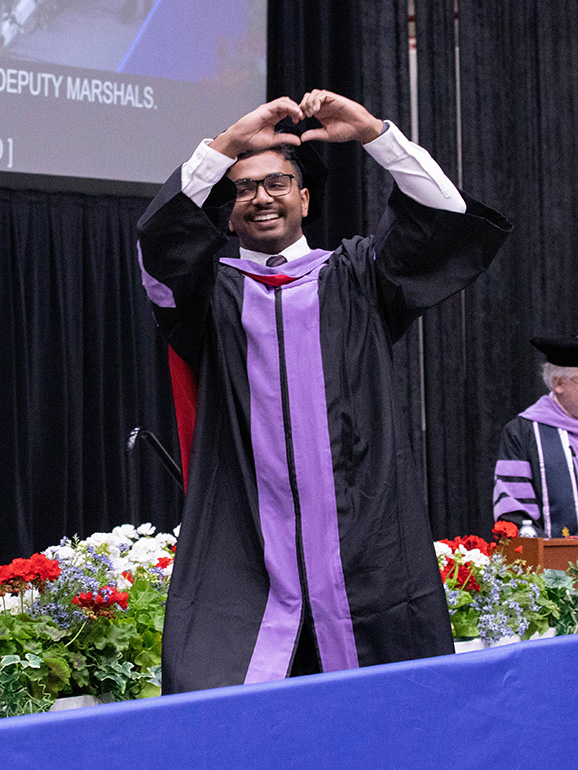 A graduate holds his hands in the air in the shape of a heart while walking across the stage at commencement.