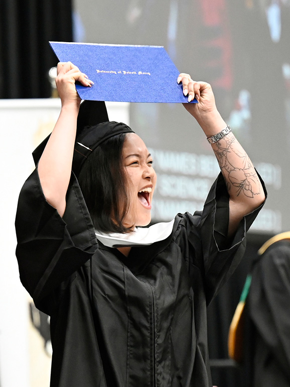A graduate holds her diploma cover in the air in celebration.