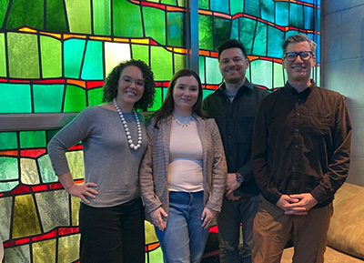 Erin Letourneau takes a photo with faculty in front of a large stained glass window.