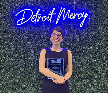 Megan Novell holds an award while standing in front of a green backdrop with a blue, neon sign that spells Detroit Mercy.