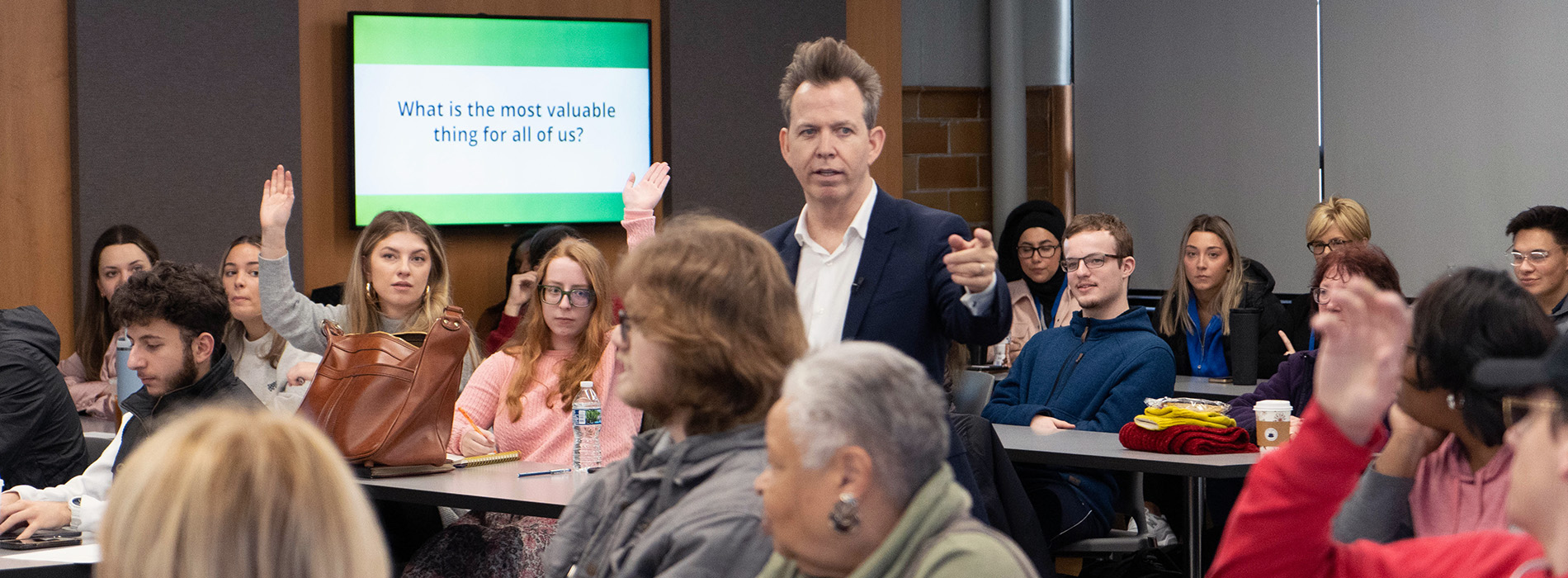 Students raise their hands as Neil Mandt stands in the center of the room during his talk on the McNichols Campus. A TV monitor in the back has text that reads "What is the most valuable thing for all of us?"