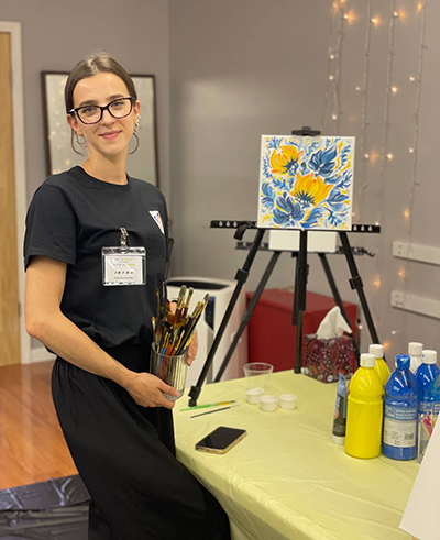 Iryna Olkhovetska poses next to her painting of blue and yellow flowers.