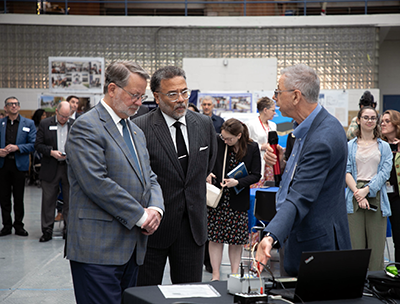 U.S. Sen. Peters and National Cyber Director Harry Coker Jr. listen and observe as a man talks about something cybersecurity related while pointing to a computer. 