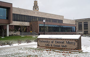 A photograph of the Student Union on Detroit Mercy's McNichols Campus during snowfall. The University of Detroit Mercy Lillie B. Kassab Mall Memorial and clocktower are visible in the foreground and background, respectively.
