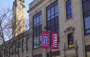 A photo of the Engineering Building near the clock tower with red banners that feature the UDM logo and World Needs Titans on it.