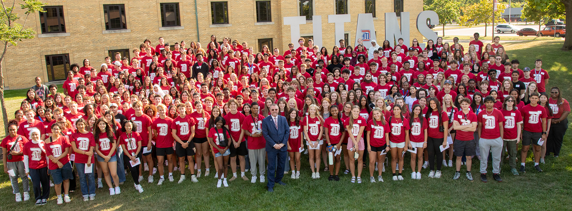 Dozens of new Titans in red shirts stand outdoors in front of the TITANS sign on the McNichols Campus.