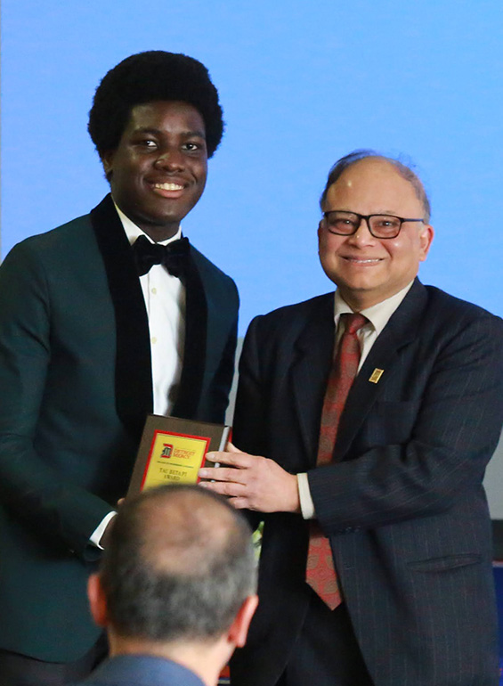 Two people dressed up stand and pose for a photo inside of the Student Union Ballroom, holding a certificate.