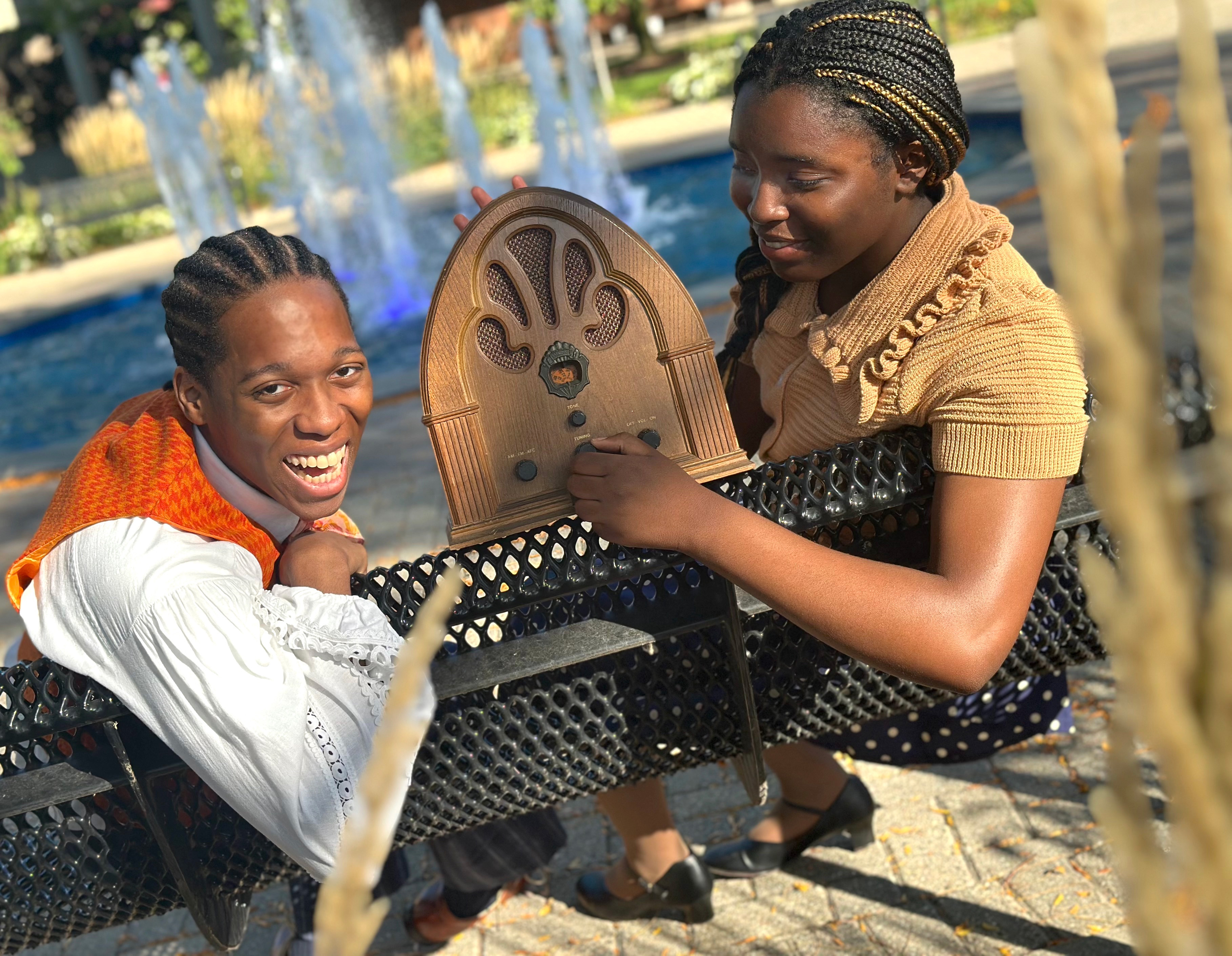 An image of two students sitting on a bench outdoors in front of a fountain, one holding an old school radio.