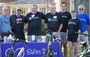 Seven people stand outdoors and pose for a photo with bicycles in front of them.
