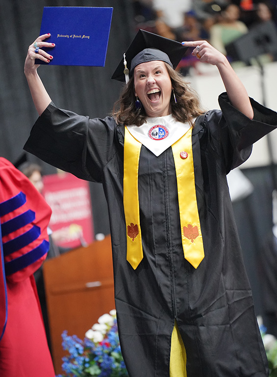 A graduate celebrates by pointing to her diploma after walking across the stage at commencement.