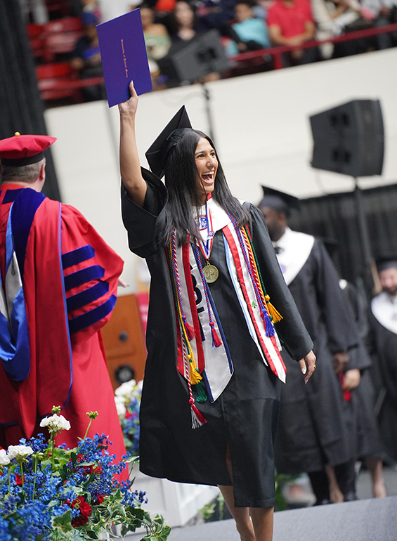 A graduate walks across the stage holding her diploma high.