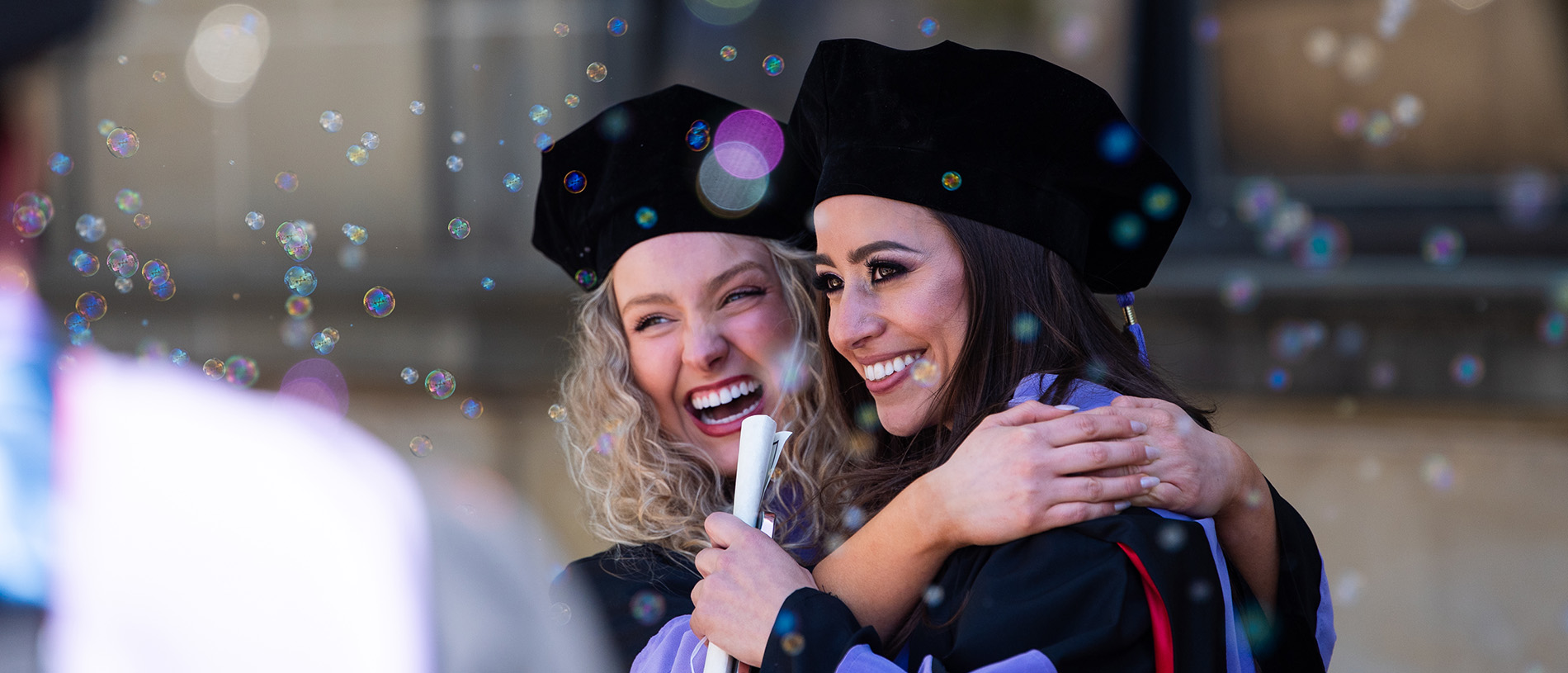 Two graduates hug following a ceremony, with bubbles floating around them.