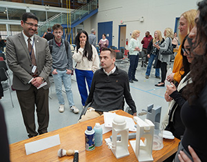 A crowd, including students from Team Michael gathers around Michael Franz and his project after the Faces on Design presentation.