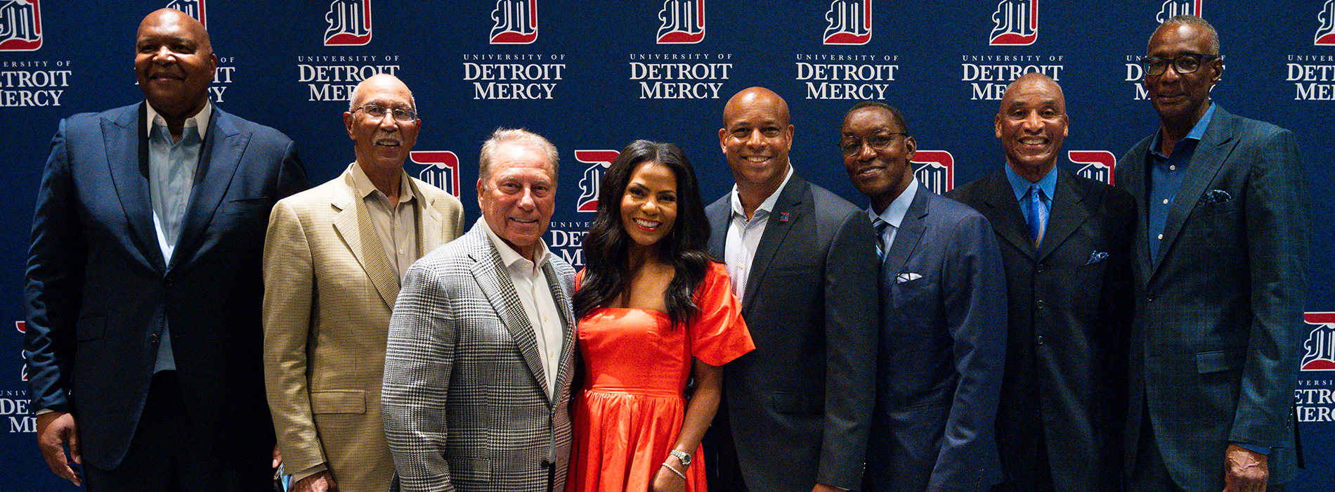 Eight people stand in front of a blue University of Detroit Mercy backdrop.
